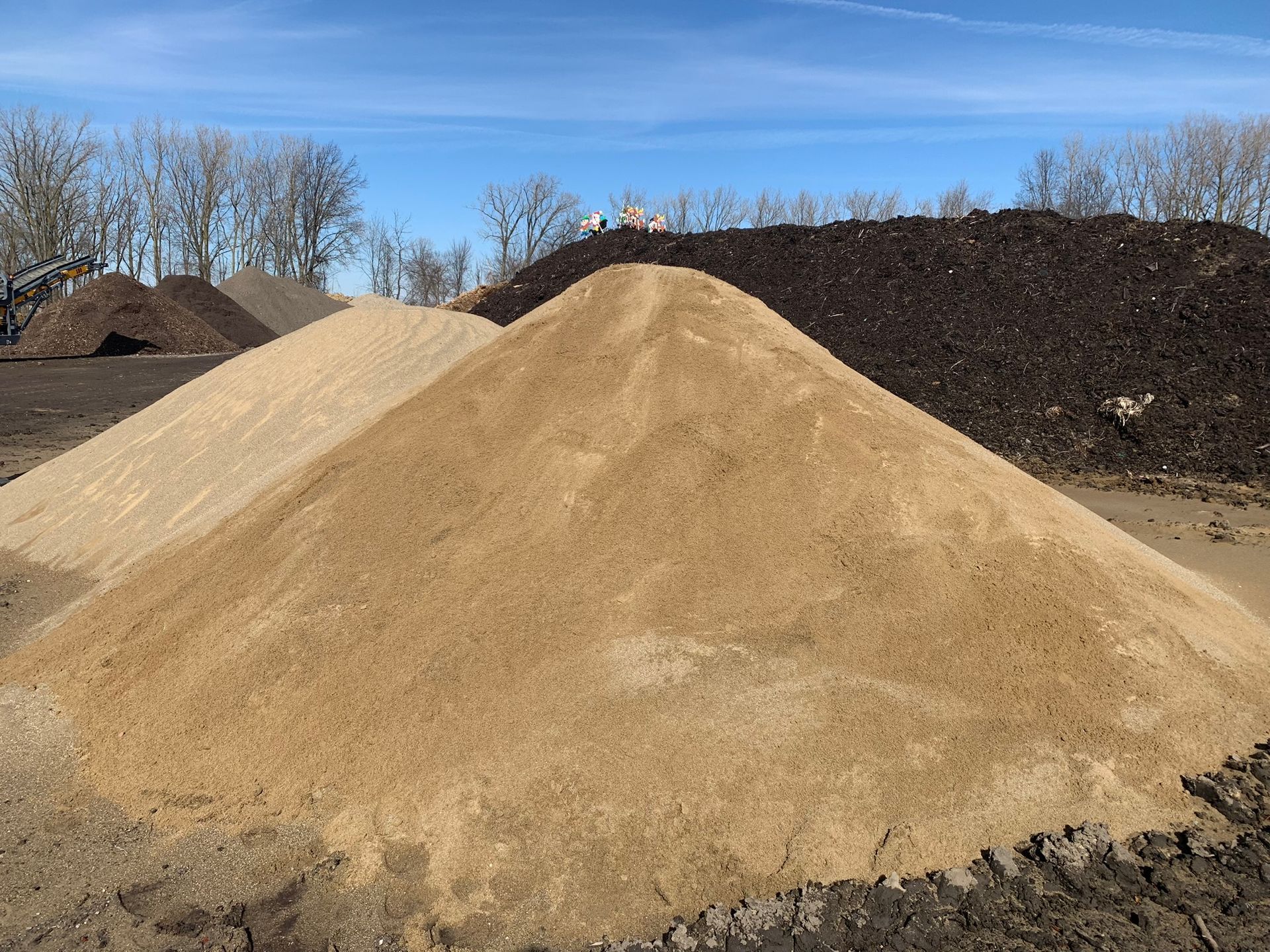 Large mounds of sand, soil, and dark mulch sit side-by-side in a storage yard under a clear blue sky.