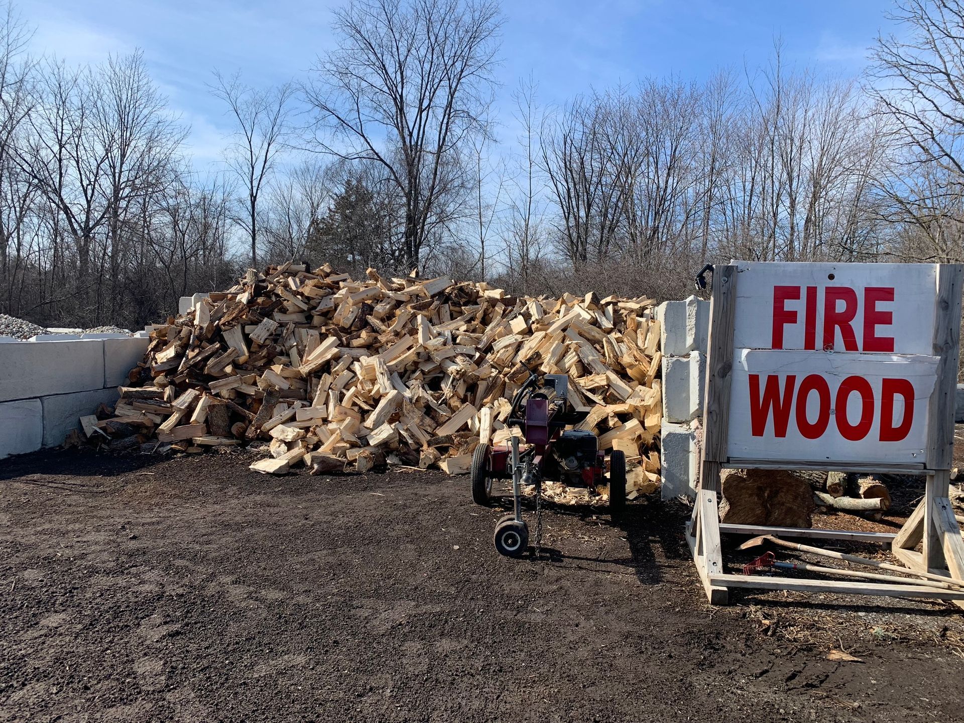 A large pile of firewood sits next to a white sign with red text that reads 