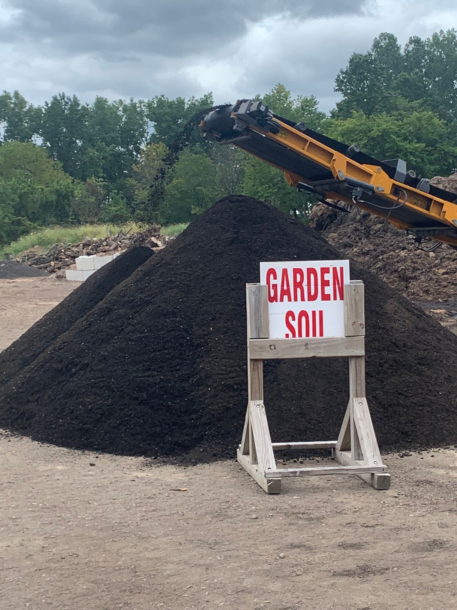 A large pile of dark garden soil sits outdoors with a wooden sign in front reading 