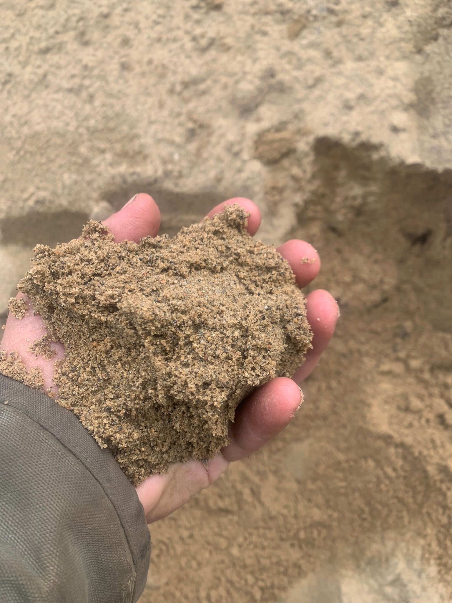 A person’s hand holds a clump of coarse, brown sandy soil against a background of a sandy excavation site.