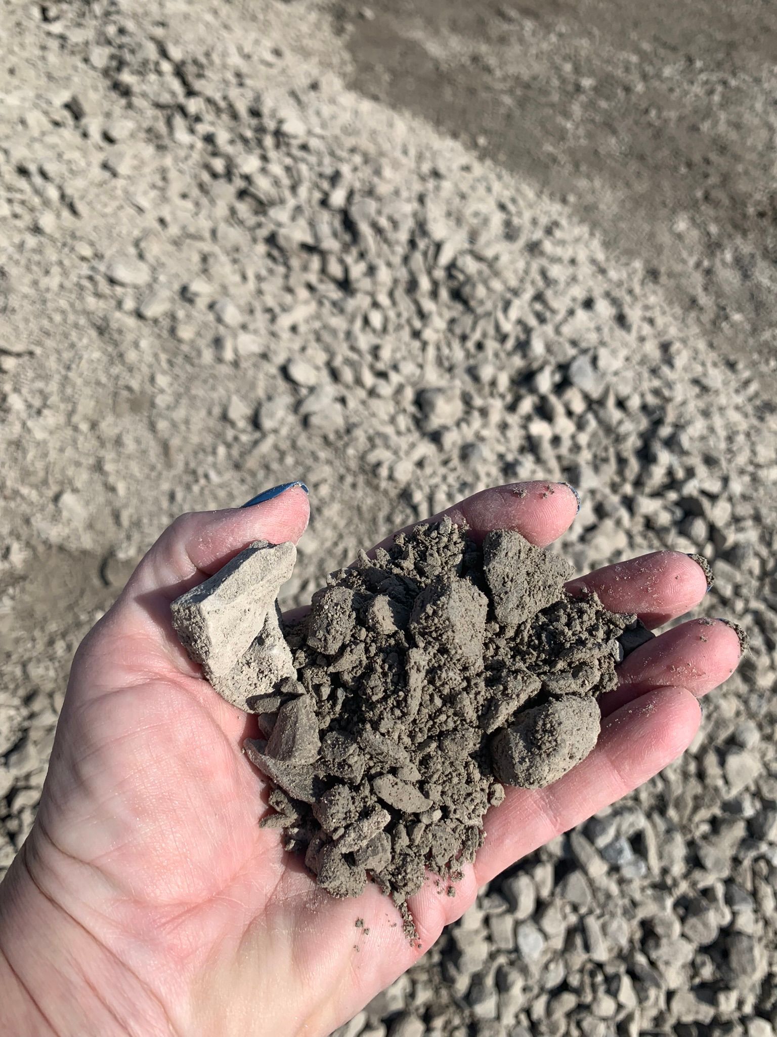 A person holds a handful of coarse, grey crushed rock and gravel against the backdrop of a large pile of similar stone.