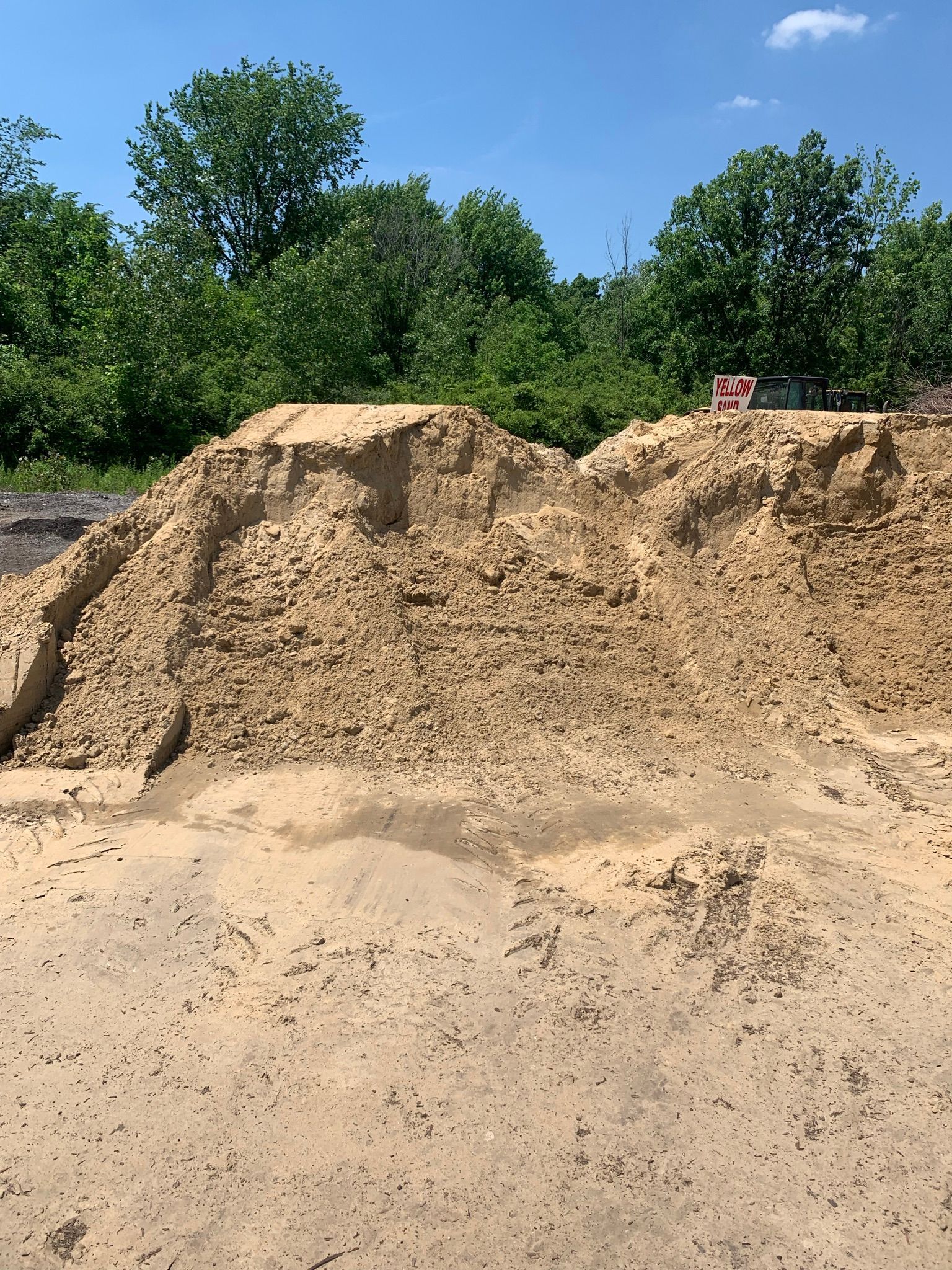 A large pile of light brown, rocky dirt sits under a bright blue sky with green trees in the background.