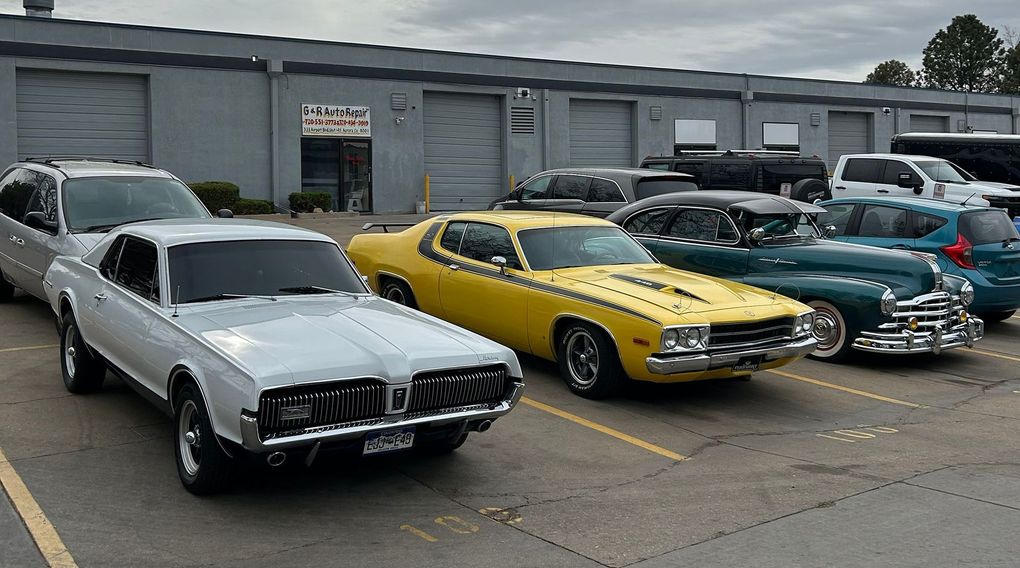 Three classic muscle cars, including a white Mercury Cougar and a yellow coupe, are parked outside a gray industrial building.