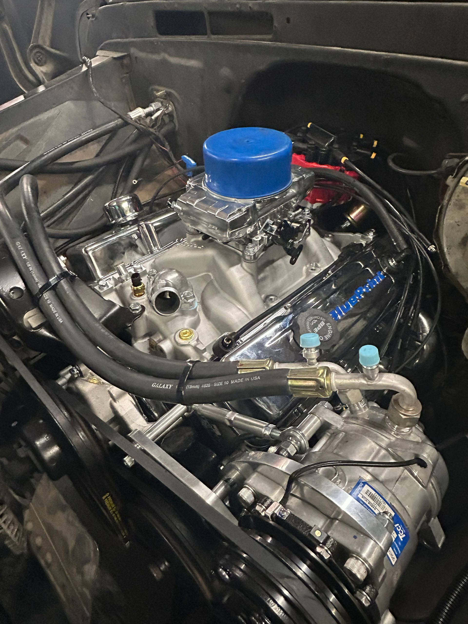A view of an open engine bay featuring a chrome intake, black valve covers, blue air cleaner top, and an AC compressor.