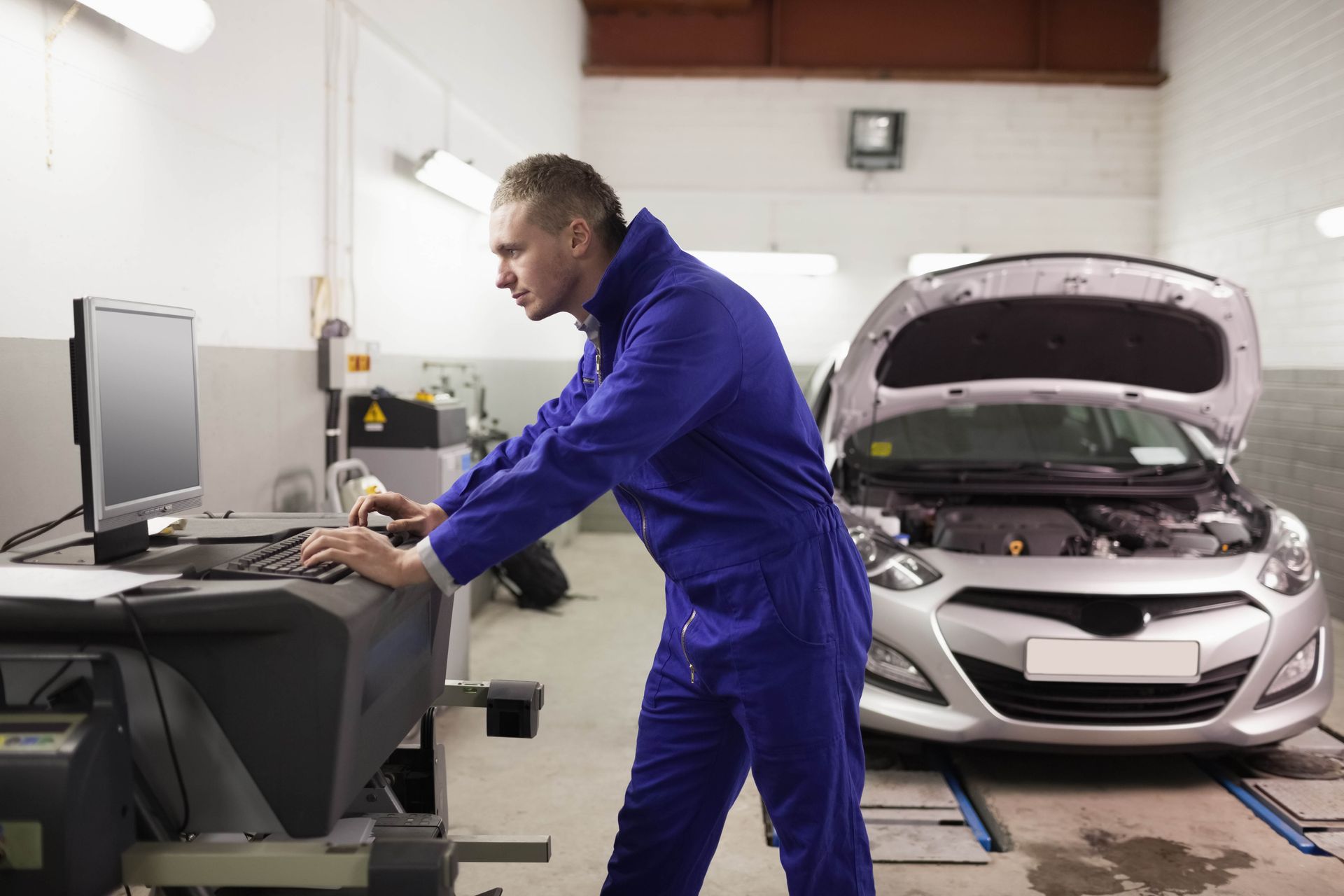 A mechanic in blue coveralls uses a computer to diagnose a silver car with its hood open in an auto repair shop.