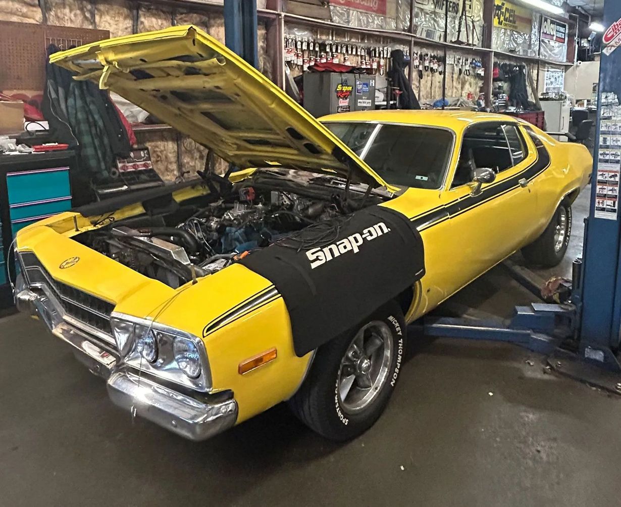 A bright yellow vintage muscle car with an open hood sits on a vehicle lift inside a mechanic's garage.