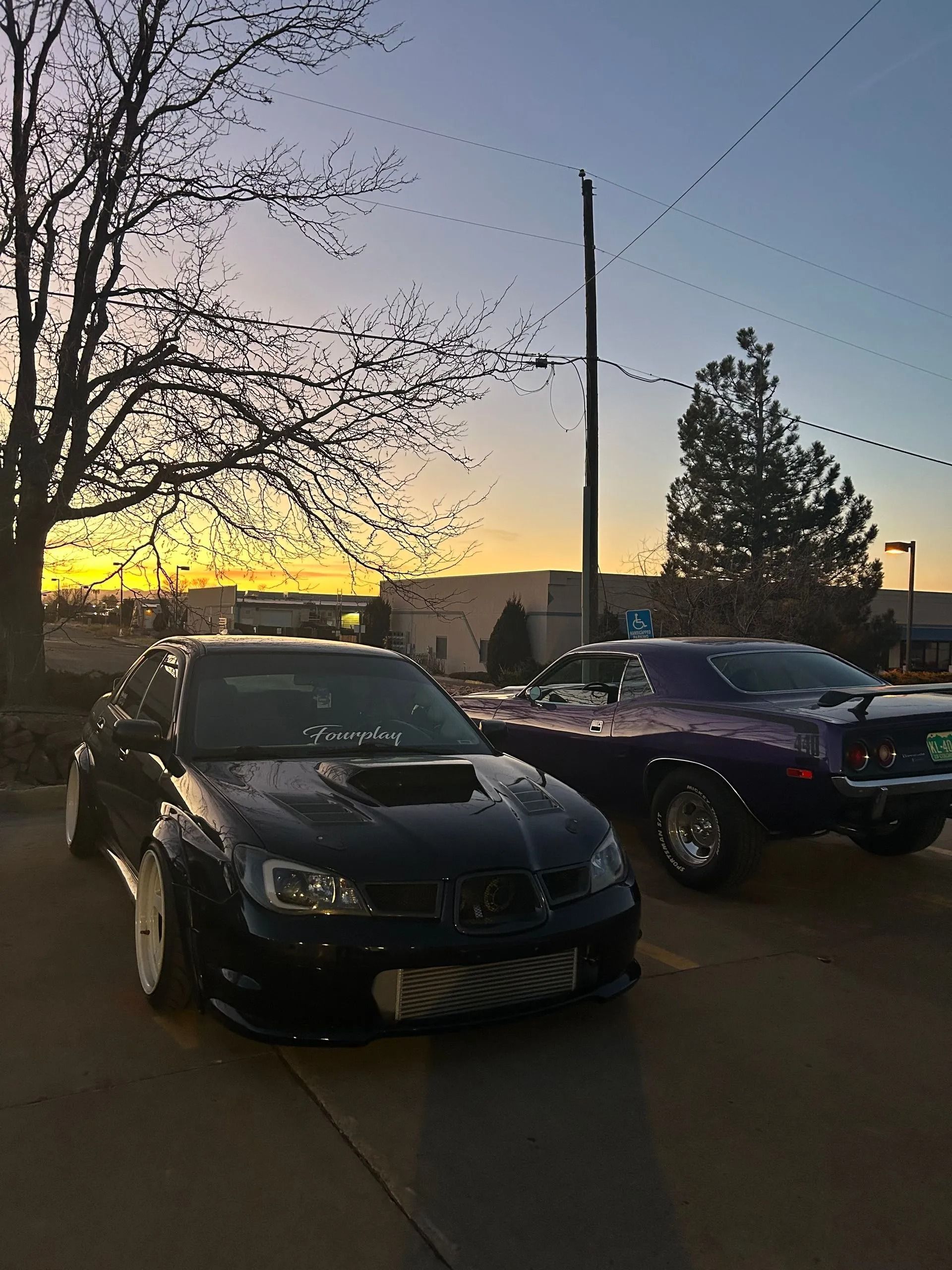 Two modified cars, a black sedan and a purple coupe, parked together on asphalt at sunset near trees and buildings.