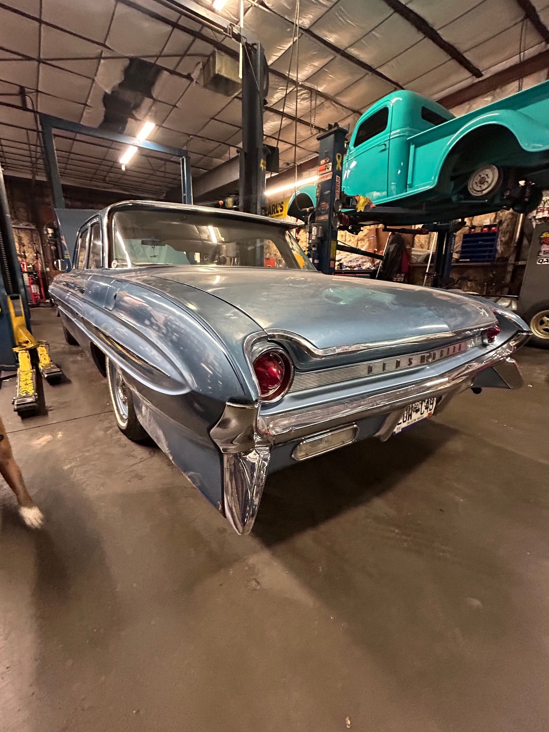 A blue vintage car in a garage, with another teal truck lifted on a service rack in the background.