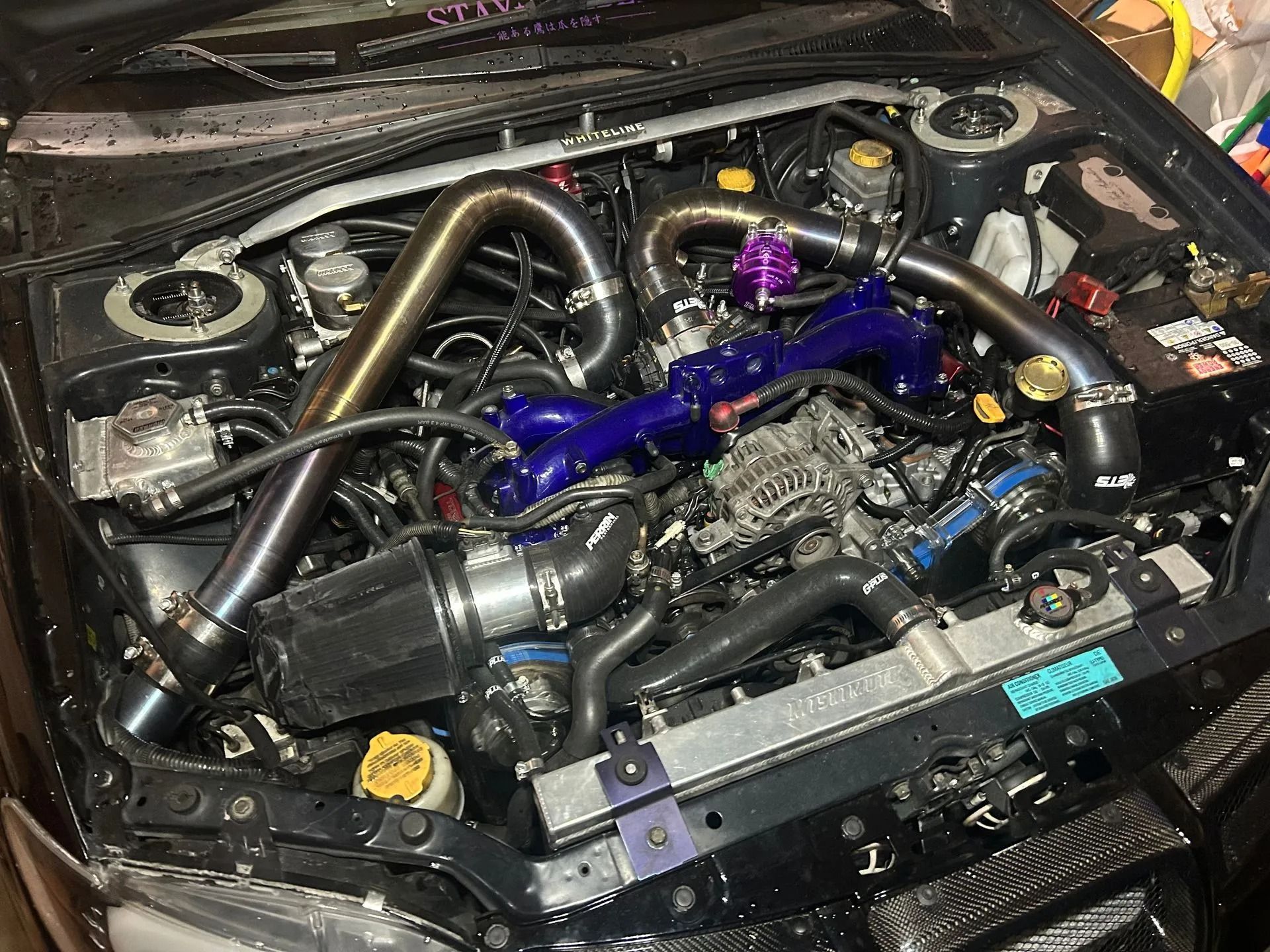 A modified turbocharged car engine bay featuring a prominent blue intake manifold and custom metal piping.