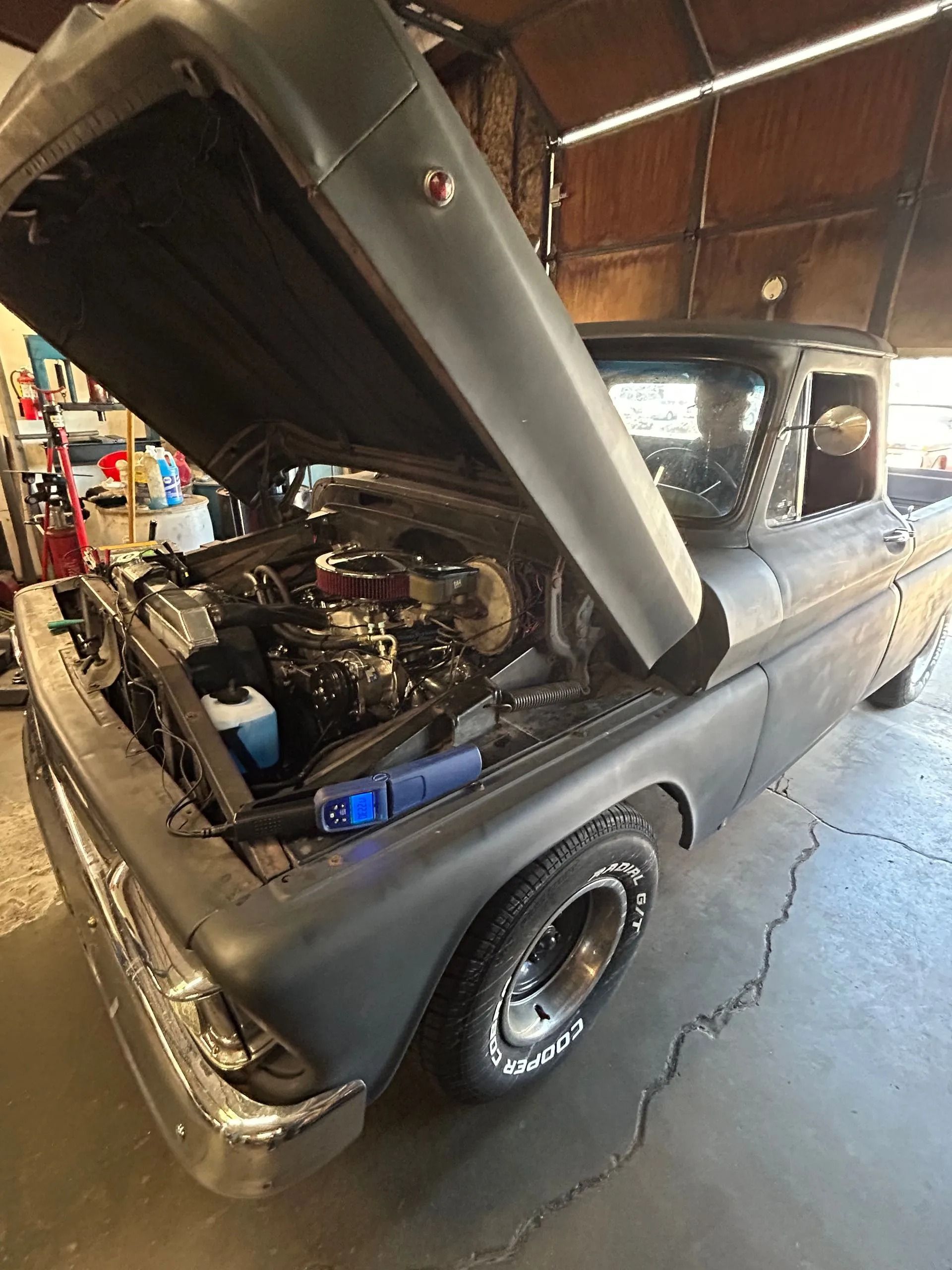 A matte black classic pickup truck parked in a garage with its hood open, revealing the engine.