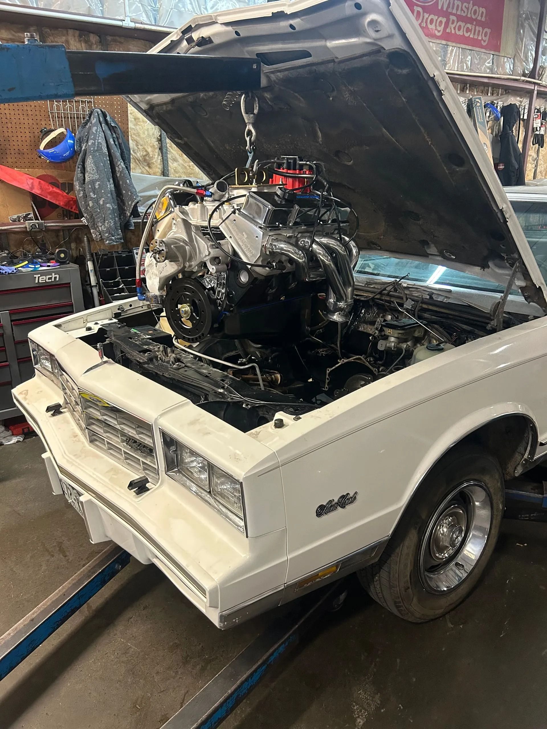 A white car with an open hood in a garage, with an engine being lowered into the engine bay by an overhead crane.
