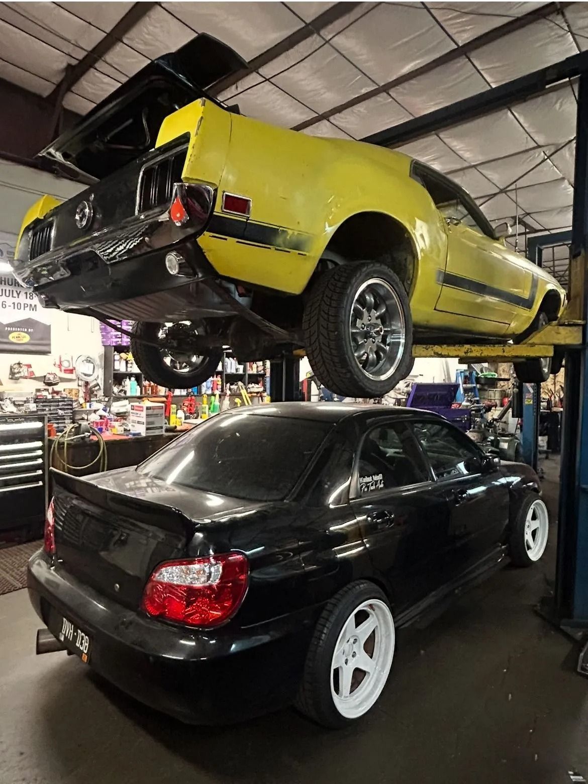 A yellow classic Ford Mustang on a car lift parked directly above a black Subaru WRX in an auto shop.