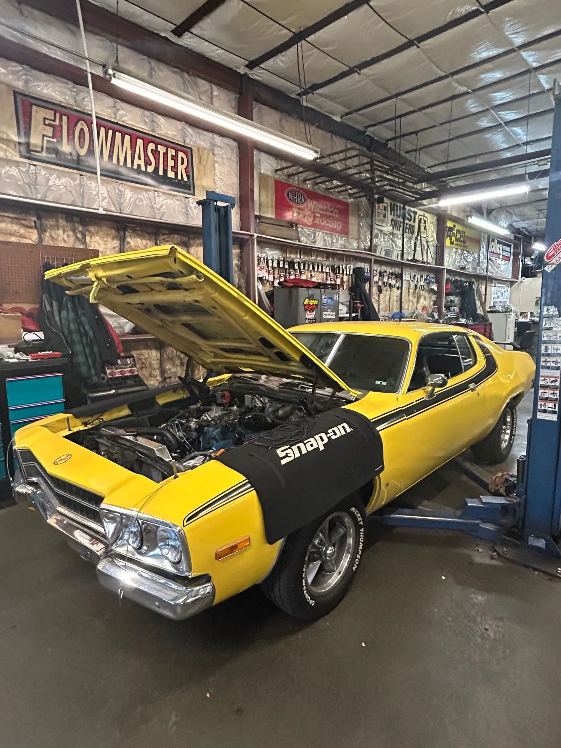 A bright yellow classic muscle car with its hood open, parked in a mechanic's garage with a Snap-on fender cover.