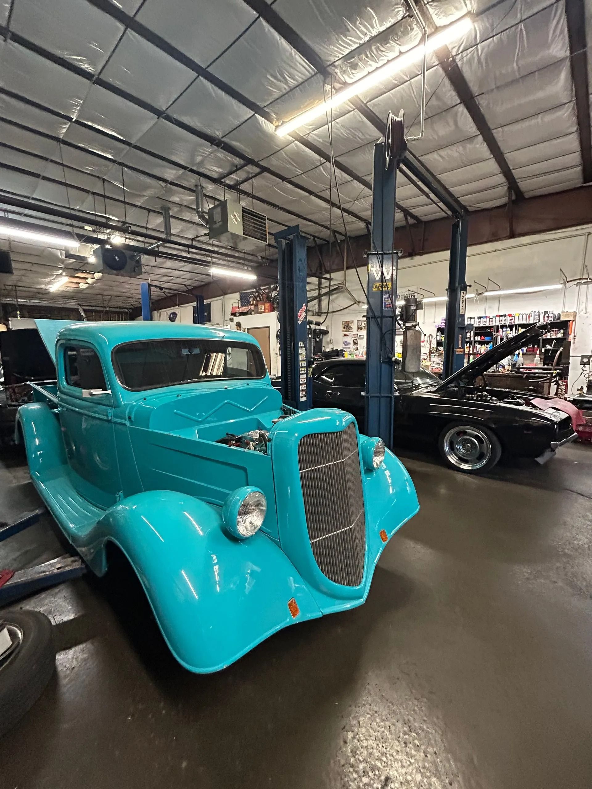 A bright turquoise vintage truck parked in a well-lit auto repair shop next to a black car under a car lift.