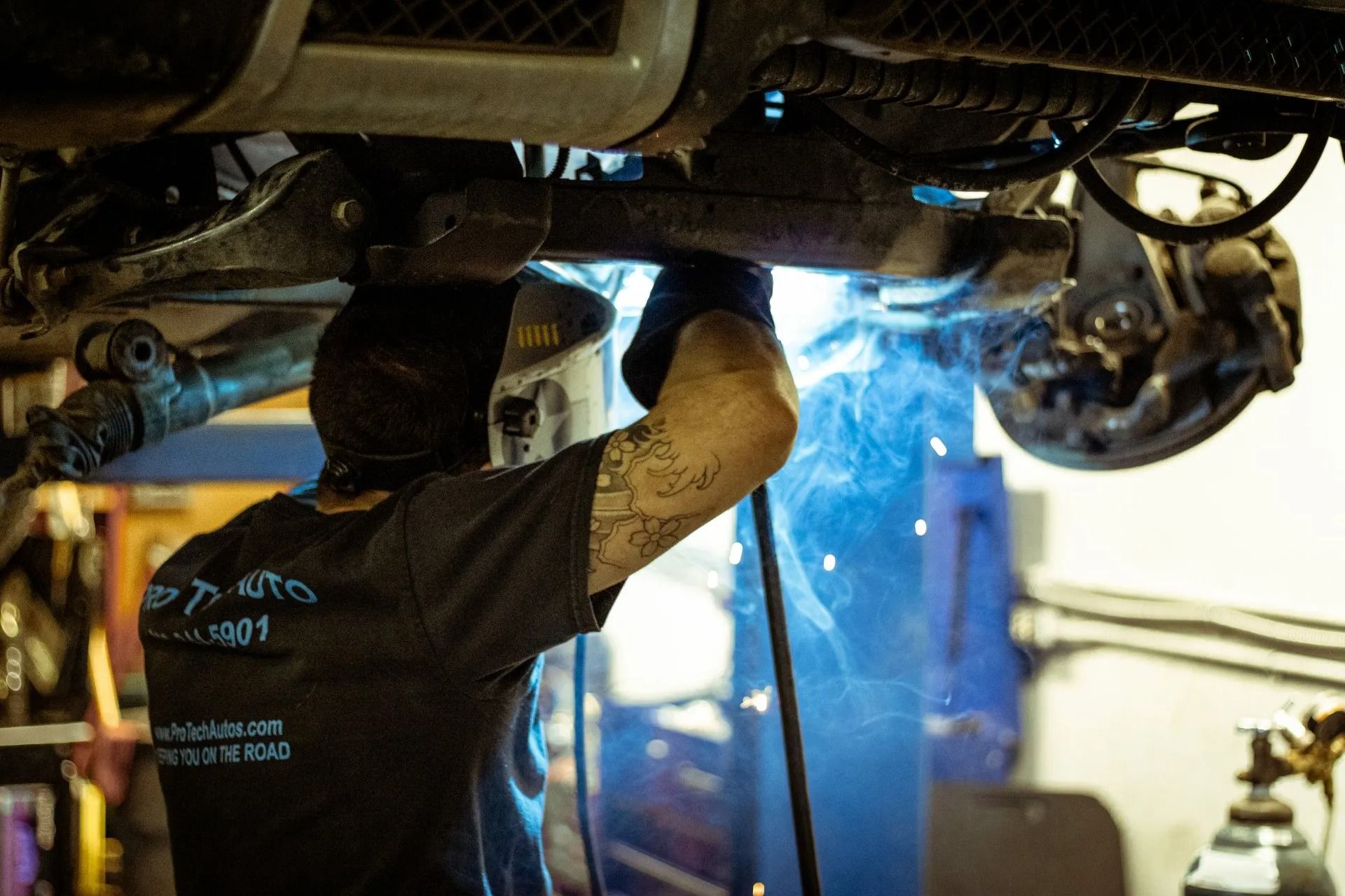 A person wearing a protective welding mask uses a torch to weld the undercarriage of a vehicle in an auto repair shop.