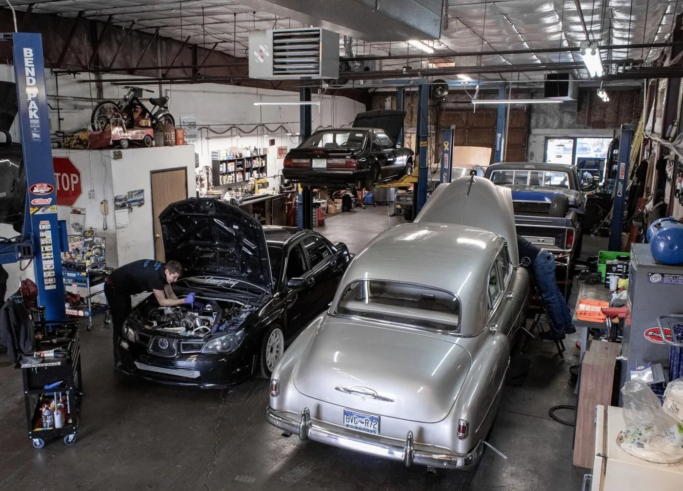 Auto repair shop with mechanics working on a black car and a silver vintage car, with another vehicle lifted on a rack.