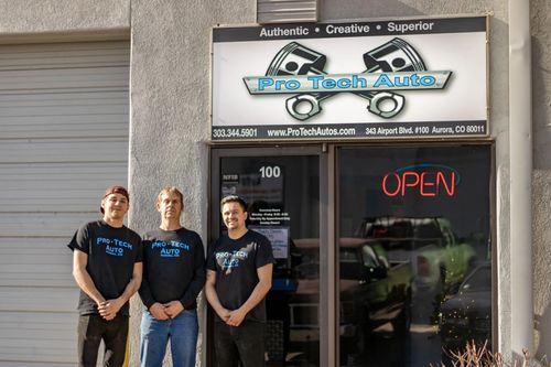 Three people standing in front of Pro Tech Auto, a business with an Open sign on its glass door.