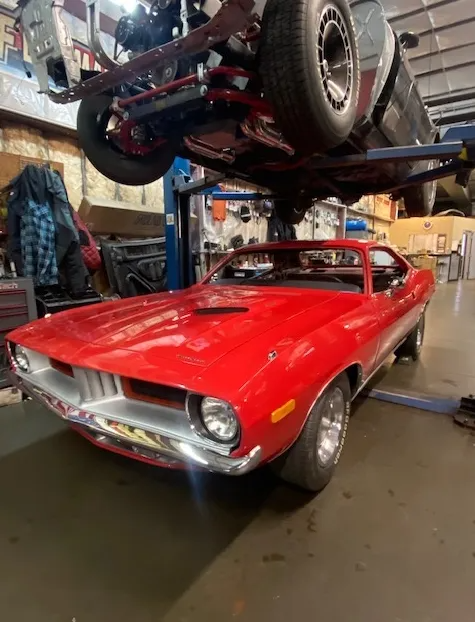 A bright red vintage Plymouth Barracuda parked inside a workshop under another vehicle raised on a car lift.