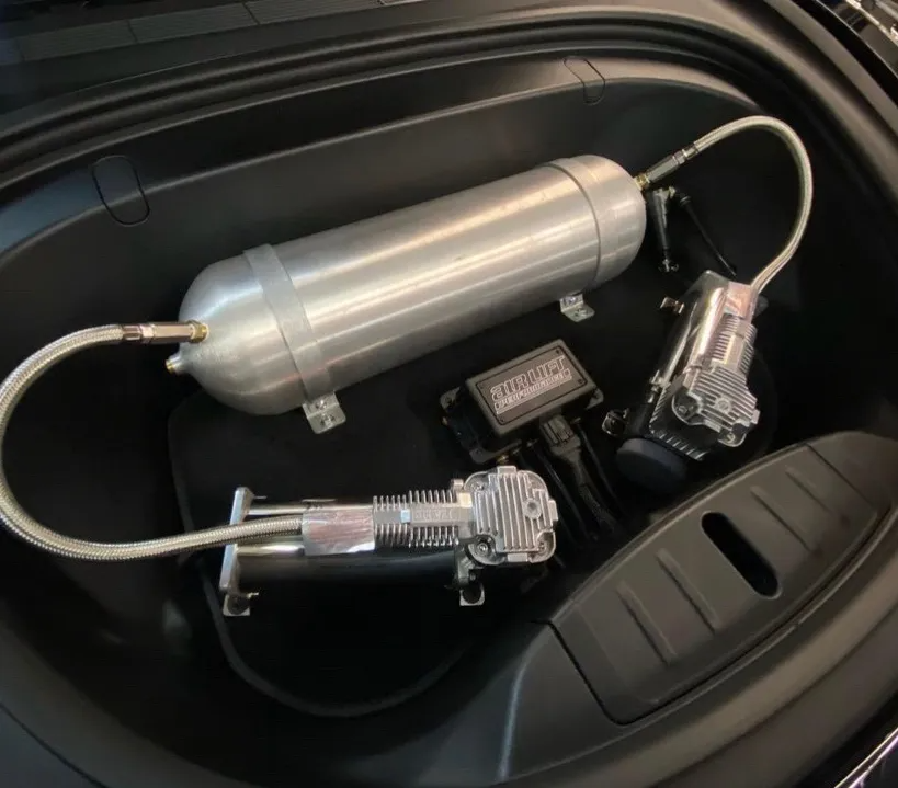 An air suspension setup featuring an aluminum tank, two chrome air compressors, and a manifold housed in a car trunk.