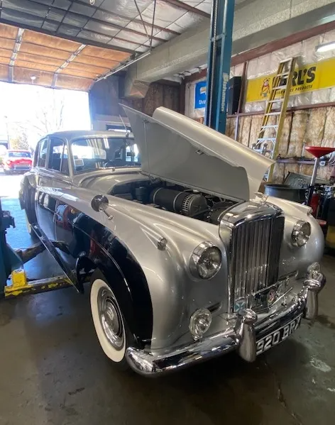 A silver and black vintage Bentley sedan parked in a repair garage with its hood raised.