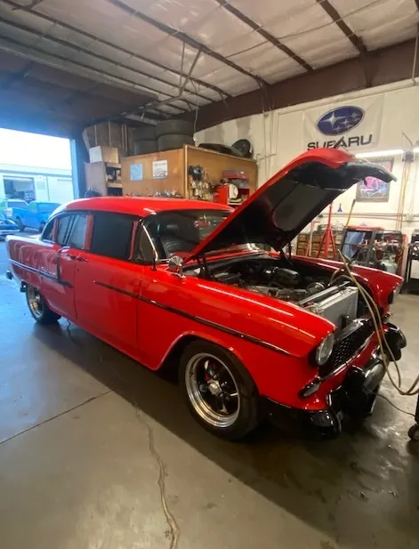 A bright red vintage sedan with its hood open, parked inside an auto repair shop with a Subaru sign in the background.