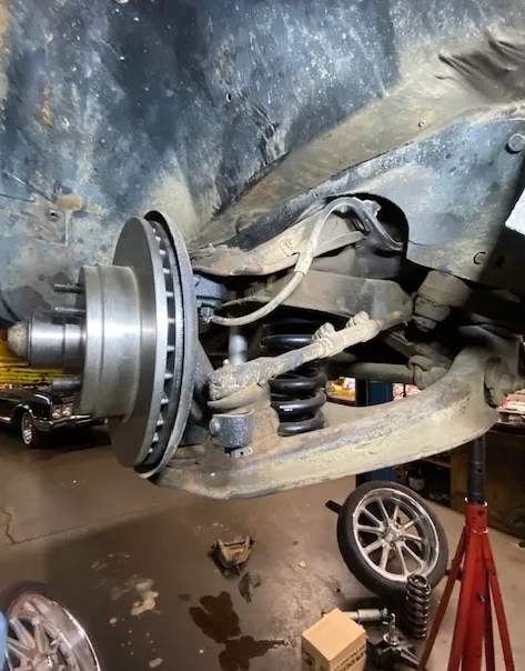 A closeup of an automotive front suspension system including the brake rotor, coil spring, and control arm in a garage.