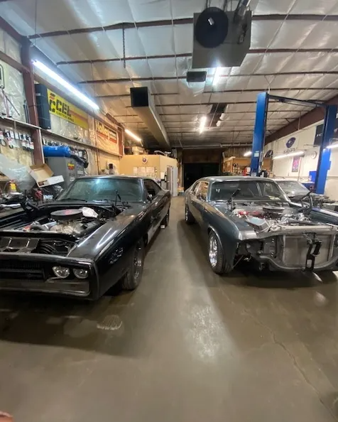 Two classic muscle cars parked side by side in a dimly lit auto repair shop with an open hood on the car on the left.