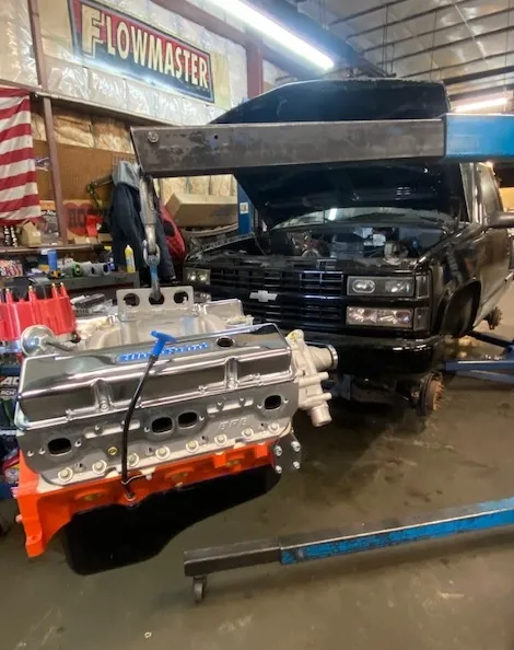An engine suspended by a hoist in a garage, positioned in front of a black Chevrolet truck with its hood open.