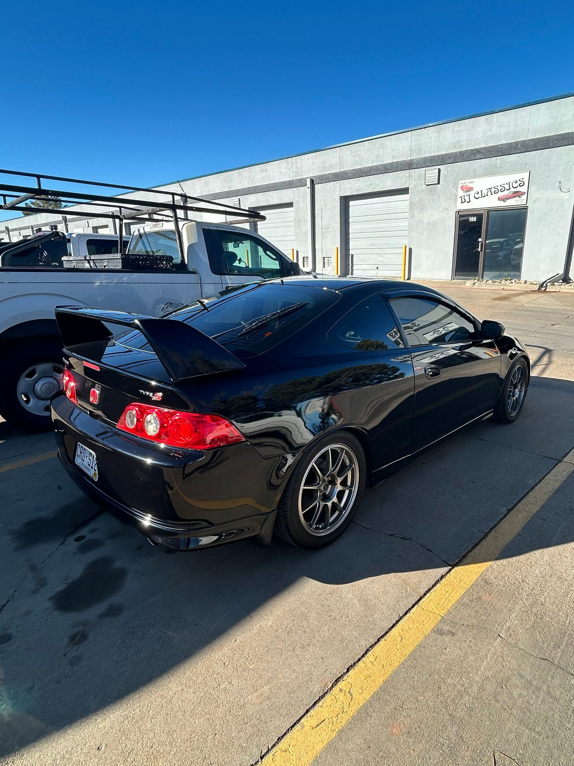 A black Acura RSX with a spoiler parked in an outdoor lot next to a white pickup truck.
