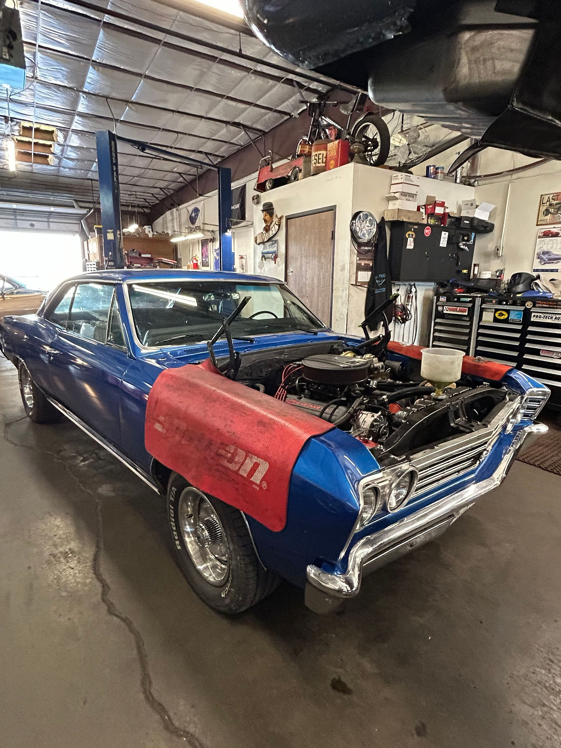 A blue vintage muscle car with an open hood inside a professional automotive repair shop.