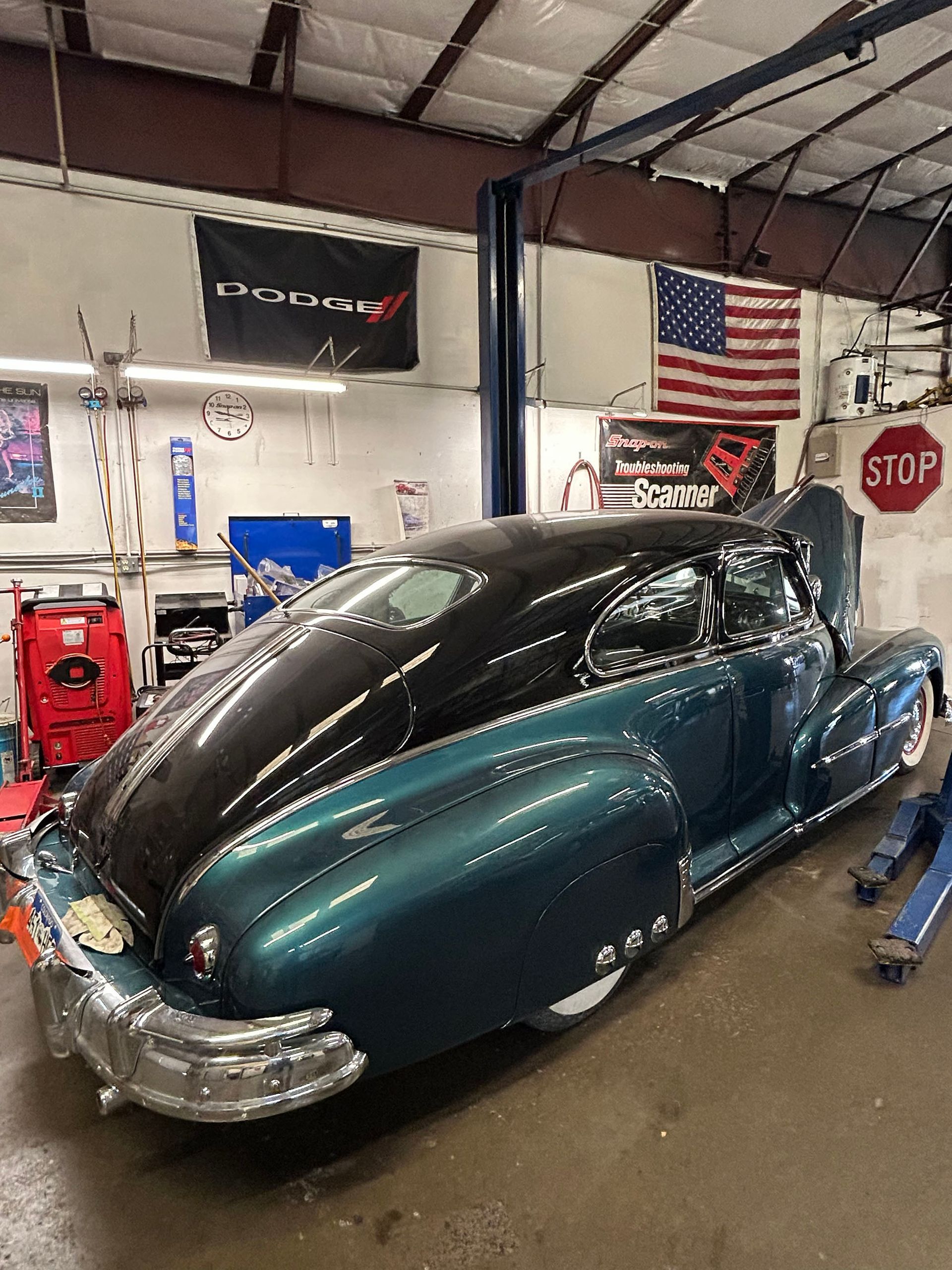 A two-tone teal and black vintage car parked inside an auto shop with an American flag displayed on the wall.