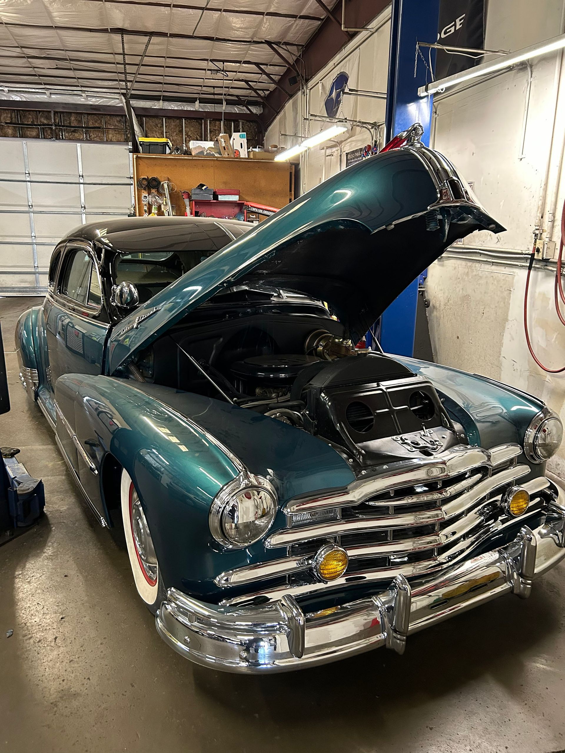 A teal vintage car with white-wall tires is parked in a garage with its hood open, revealing the engine.