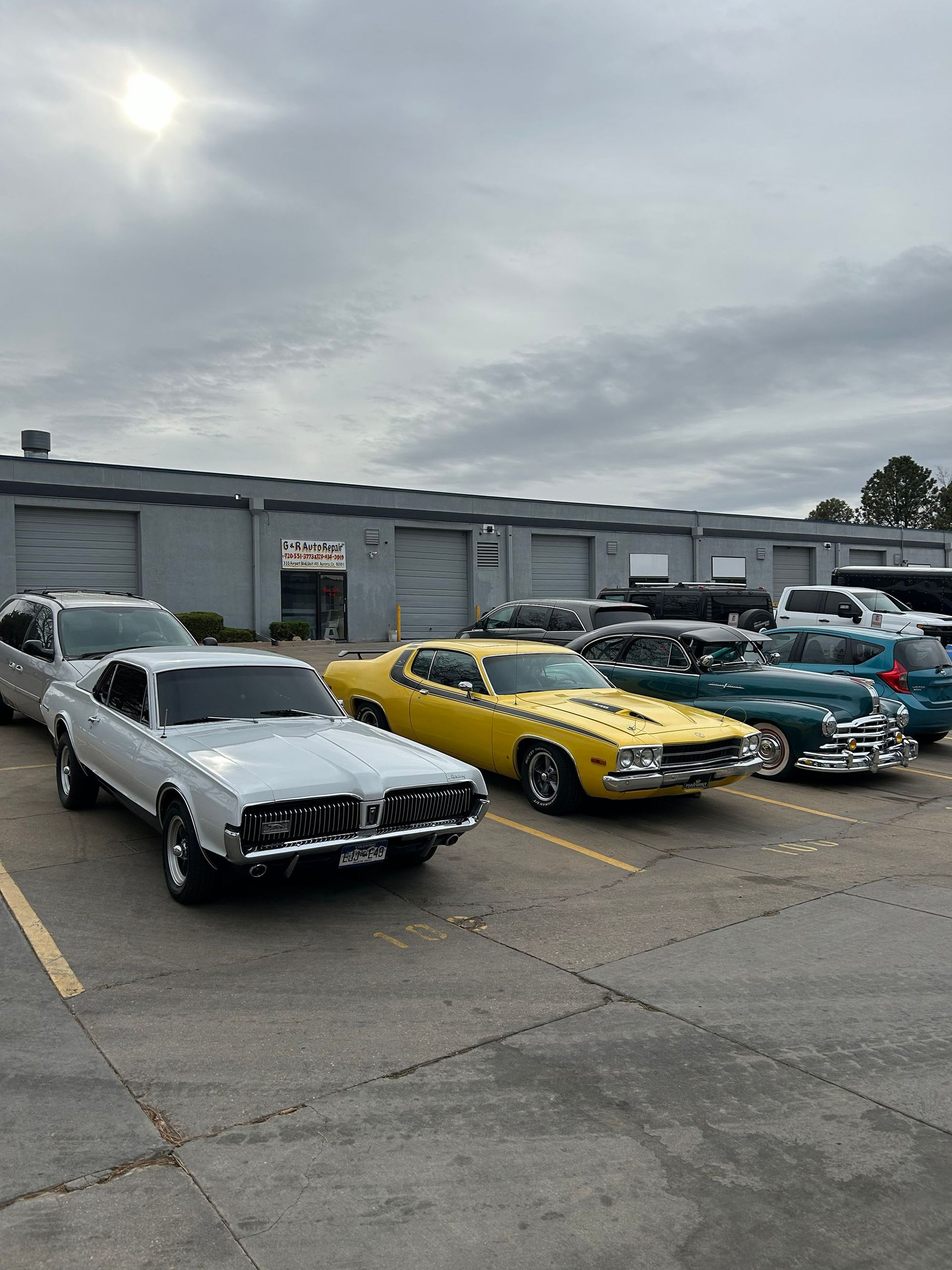 A line of classic vintage cars, including a white Mercury Cougar and a yellow muscle car, parked outside a warehouse.
