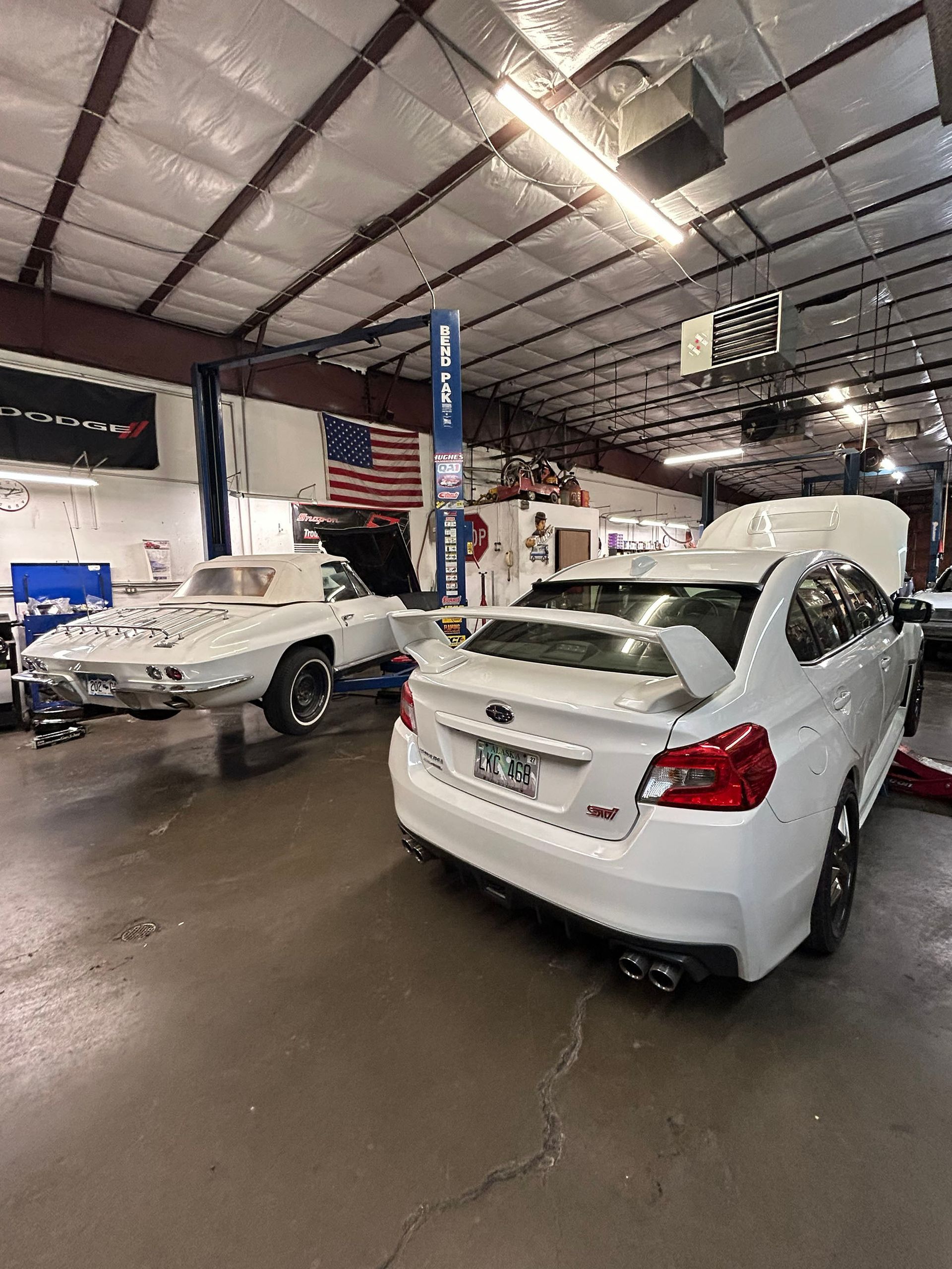 A white Subaru WRX STI parked in an auto repair shop next to a classic white convertible Corvette on a lift.