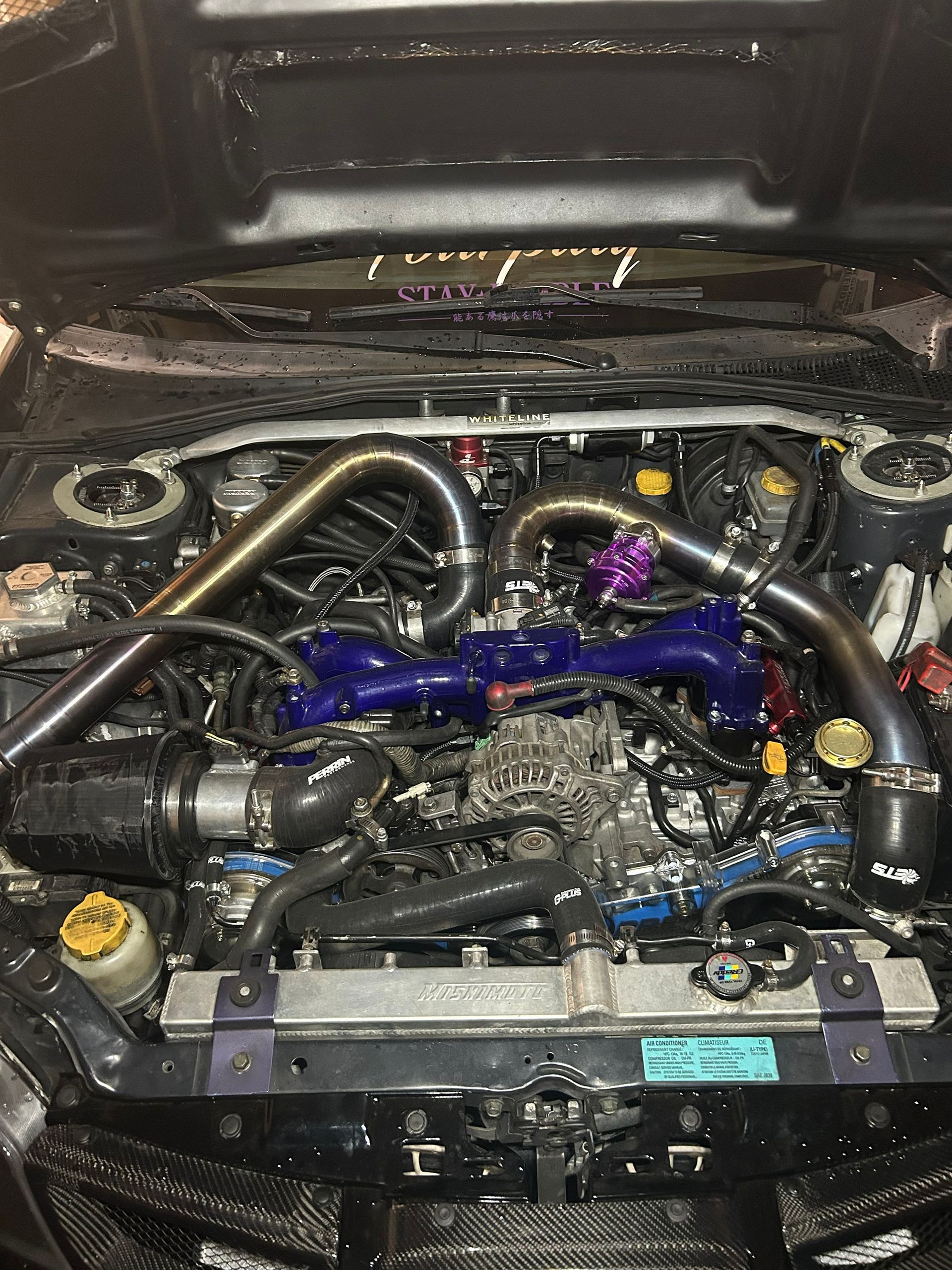 The open hood of a modified car engine bay with a prominent blue intake manifold and polished metal piping.