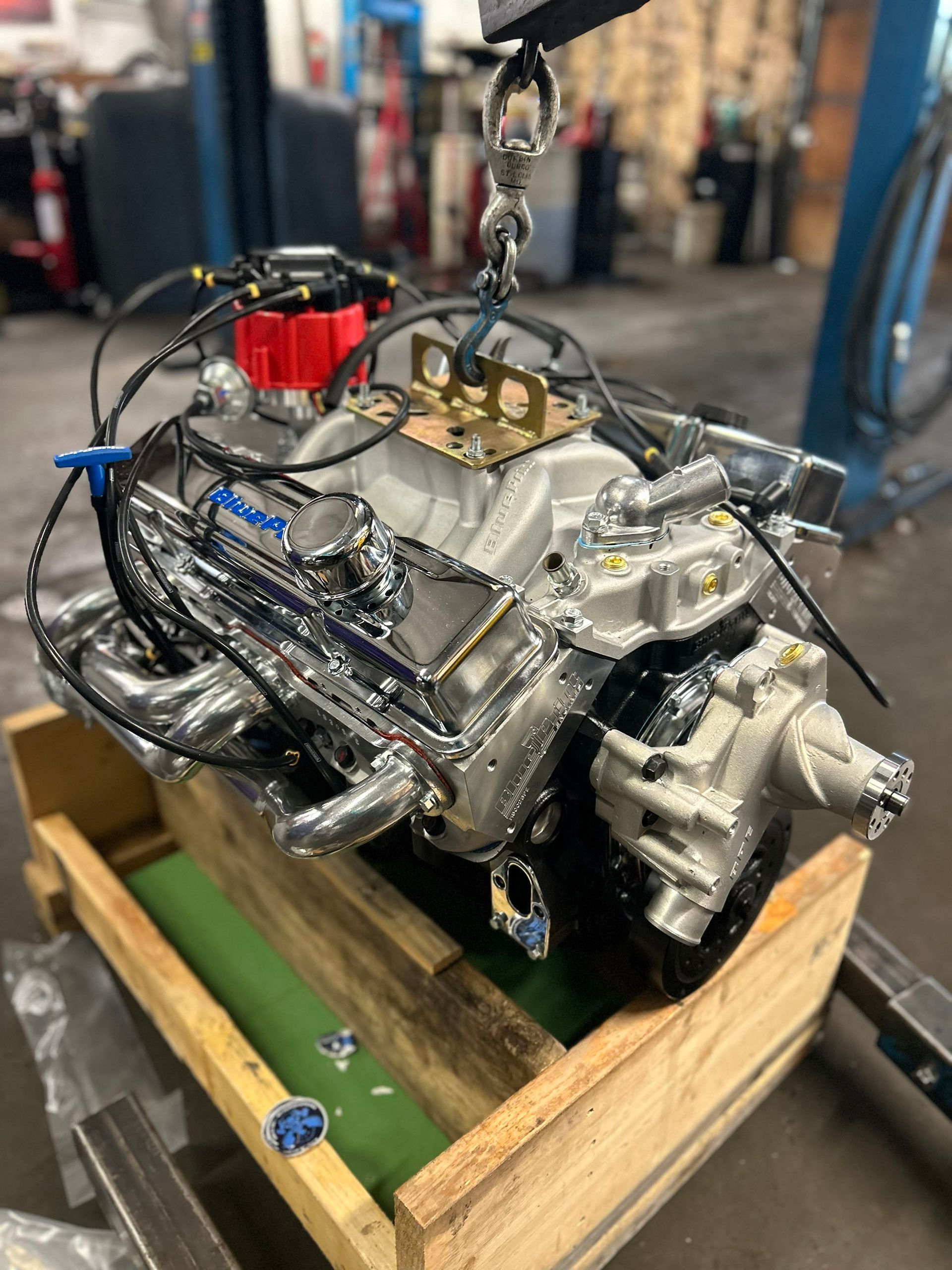 A polished V8 engine with chrome valve covers and a red ignition distributor hanging from a chain in a workshop.