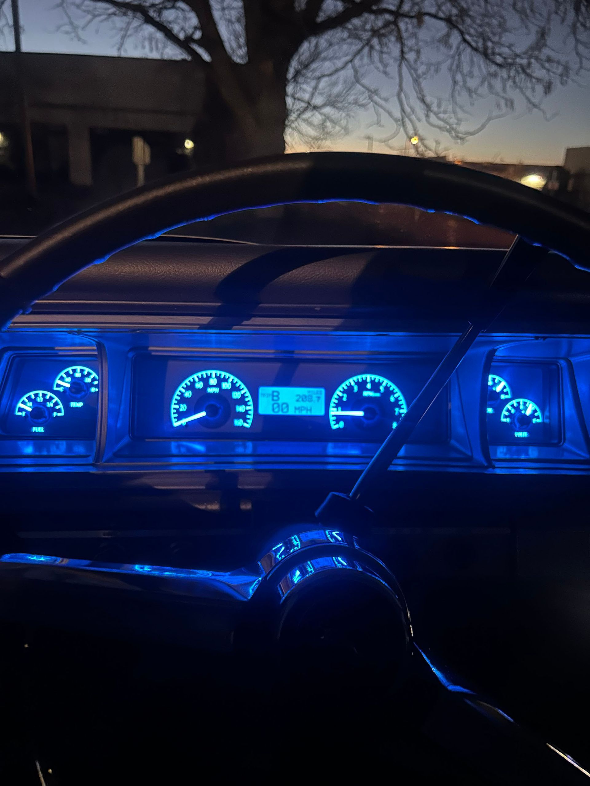 Car dashboard with glowing blue analog gauges and a digital display at dusk.