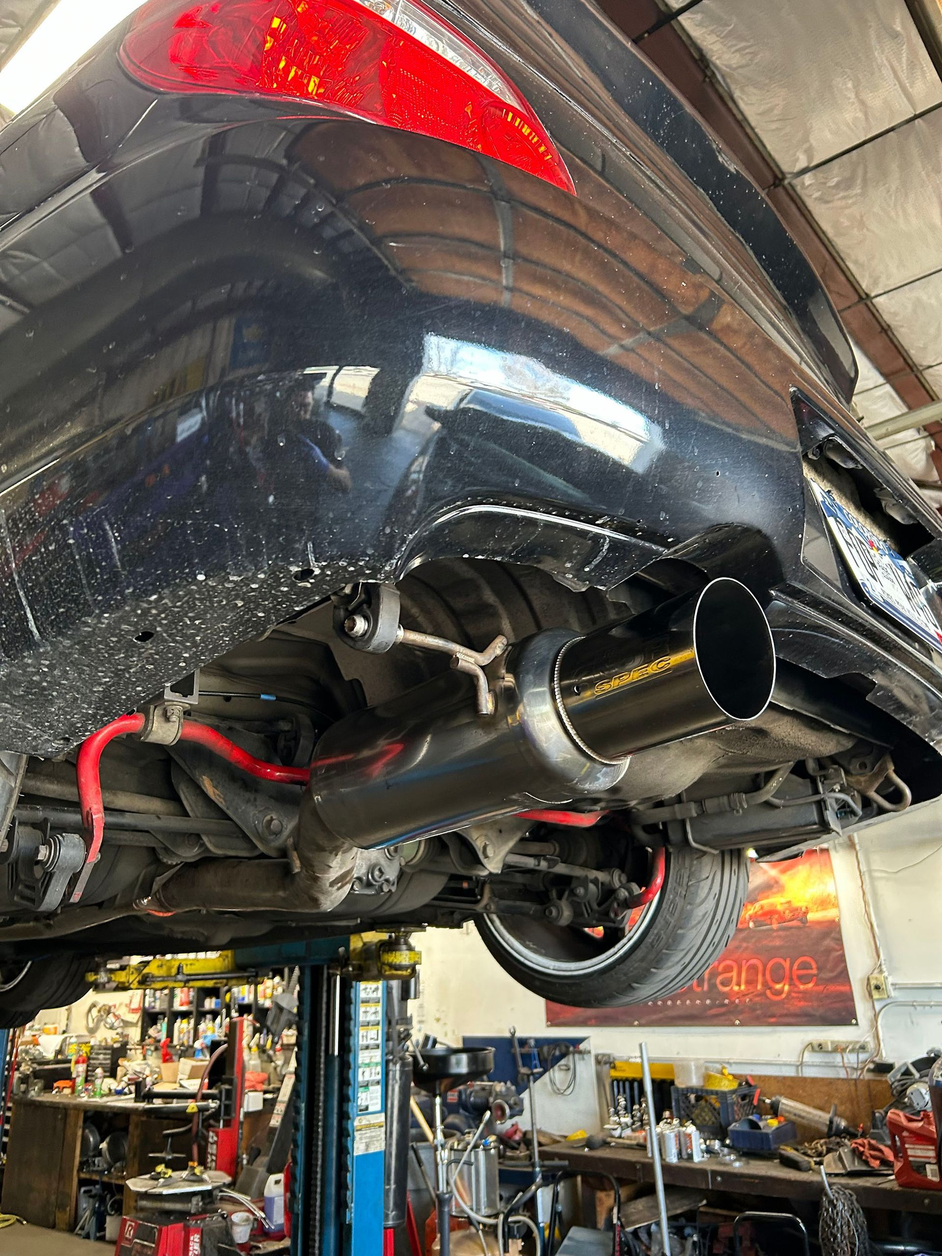 Underside of a black car on a mechanic's lift, showcasing a large, aftermarket exhaust pipe and red sway bar.