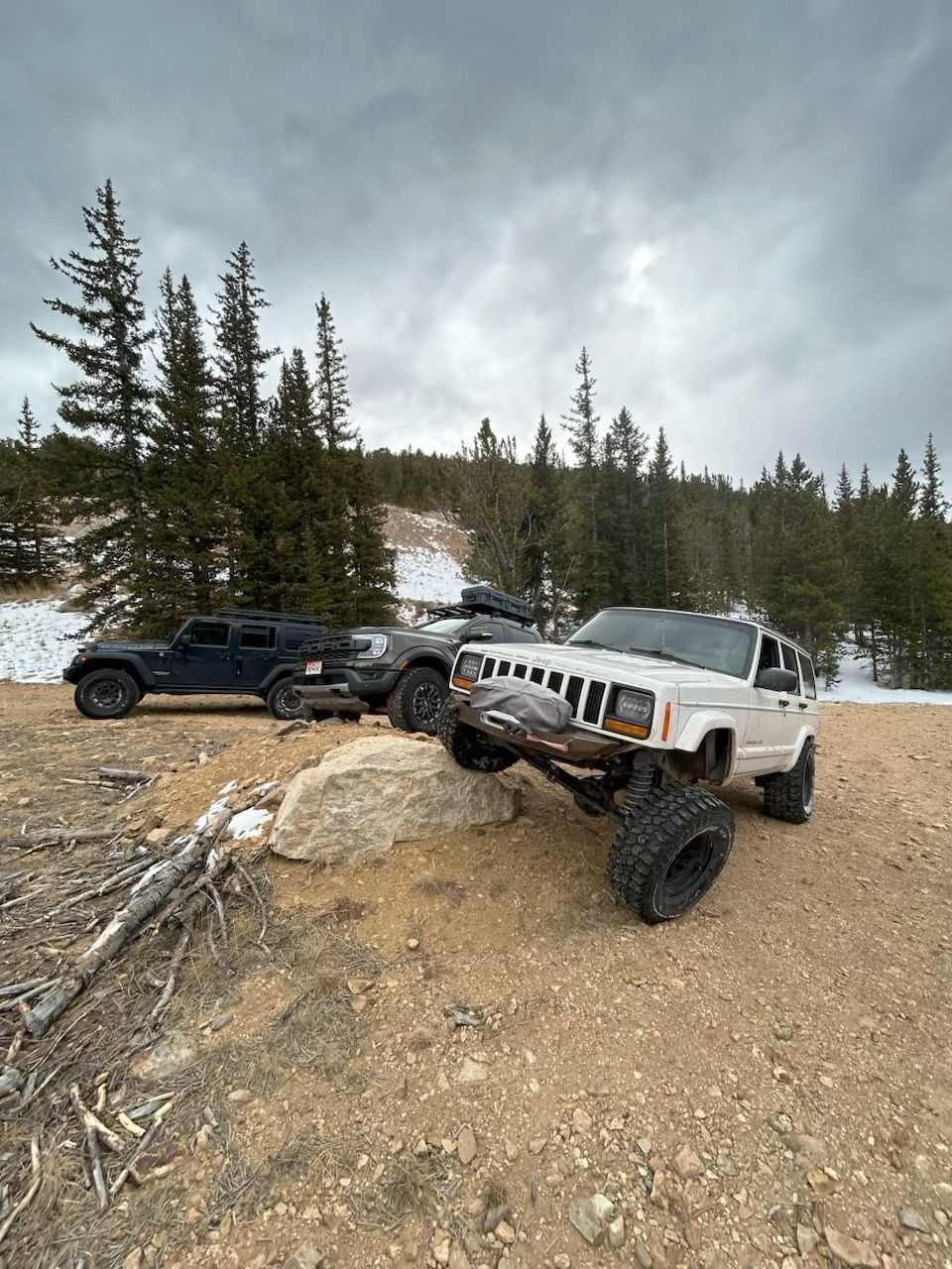 A white Jeep flexes its front suspension over a large rock near two other parked vehicles in a snowy, wooded area.