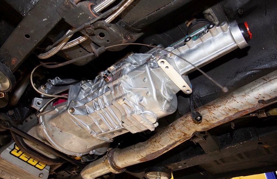 Under-vehicle view of a clean, silver automotive manual transmission mounted to a chassis with metal framing and pipes.