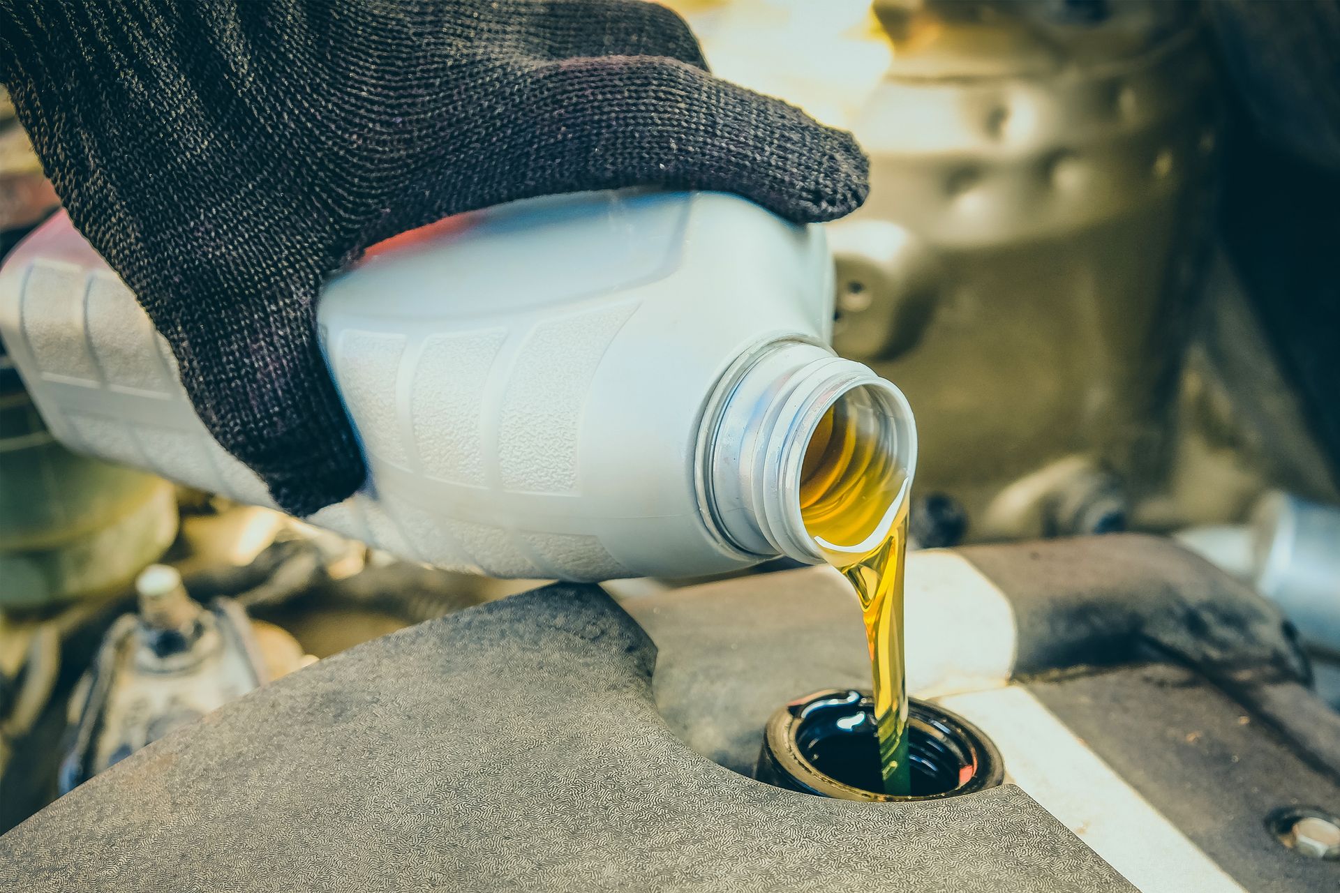 A gloved hand pouring golden engine oil from a plastic bottle into a car engine.