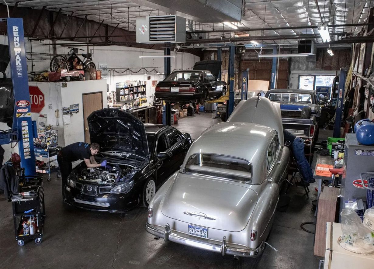 A busy auto repair shop with mechanics working on a black car and a vintage silver car, with others on vehicle lifts.