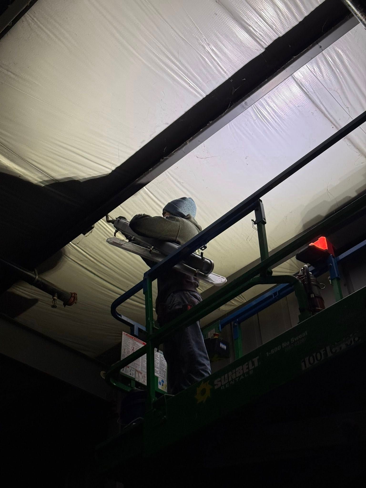 A person wearing a beanie stands on a green scissor lift, working on the ceiling of an indoor structure.