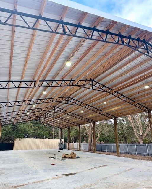 Large open-sided steel-roof pavilion with a concrete floor, set among trees and fencing.