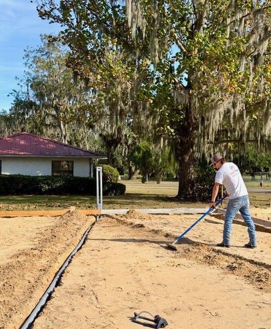 A person in a white shirt and jeans uses a rake to level sand at a construction site near a tree-lined yard.