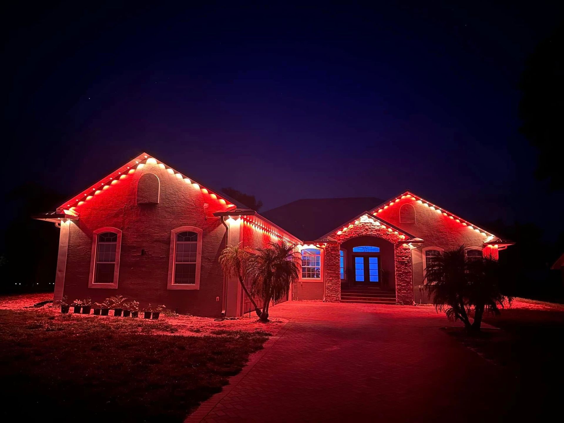 A house at night decorated with vibrant red holiday lights along the roofline, with red light cast across the front yard.