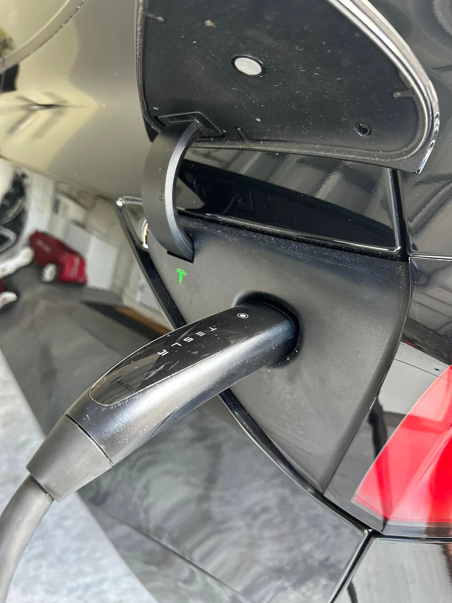 A black electric vehicle charging port with a plugged-in charger, featuring a small, glowing green status light.