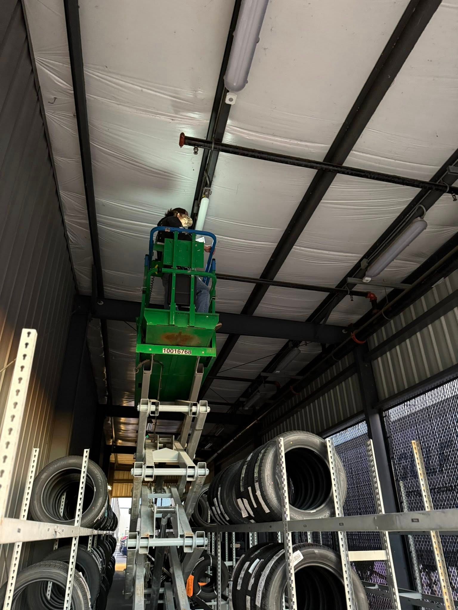 A person on a green scissor lift working on lights installed on the ceiling of a warehouse filled with stacks of tires.