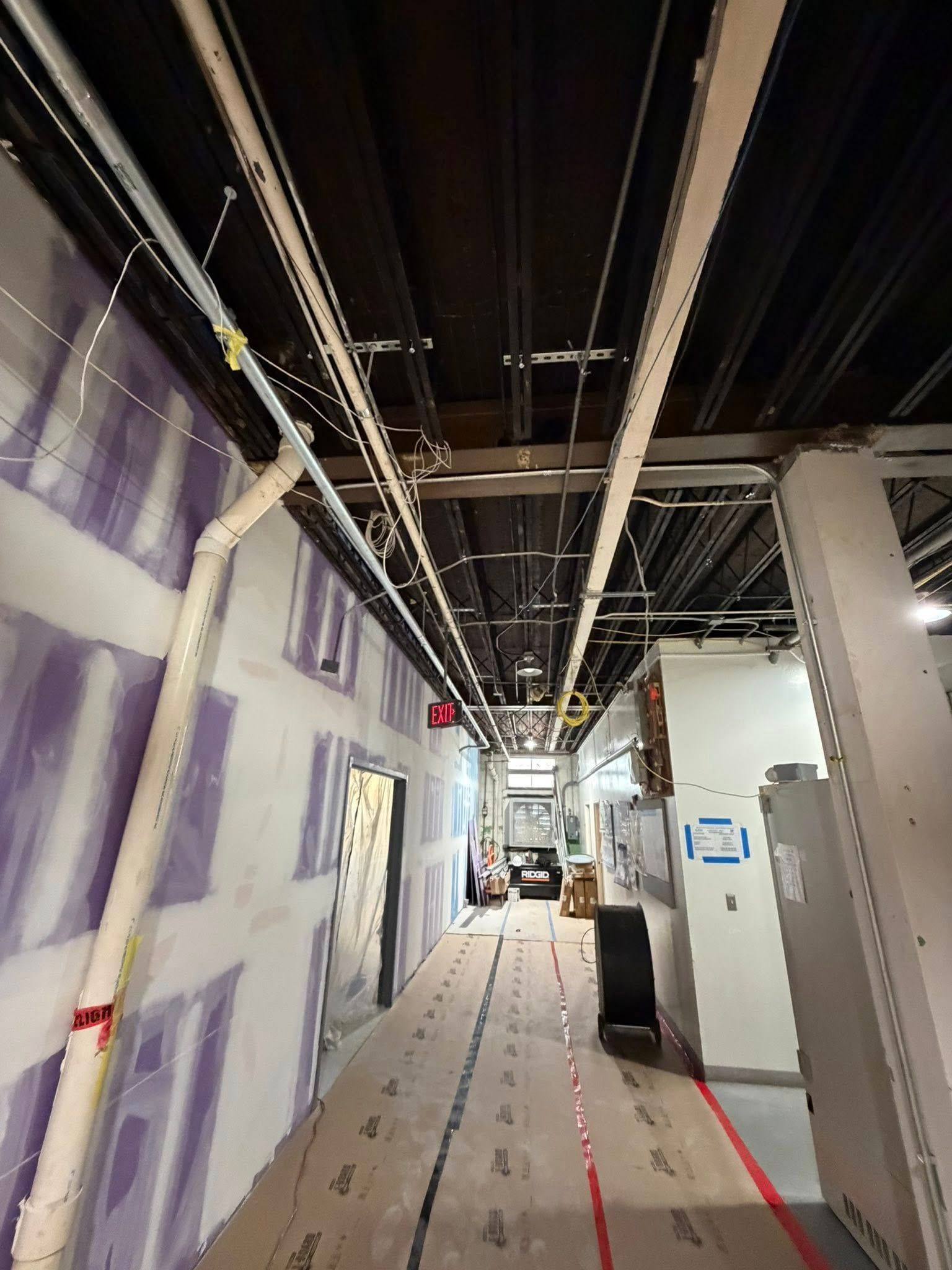 An interior construction corridor with exposed ceiling pipes, white drywall panels, and a protective floor covering.