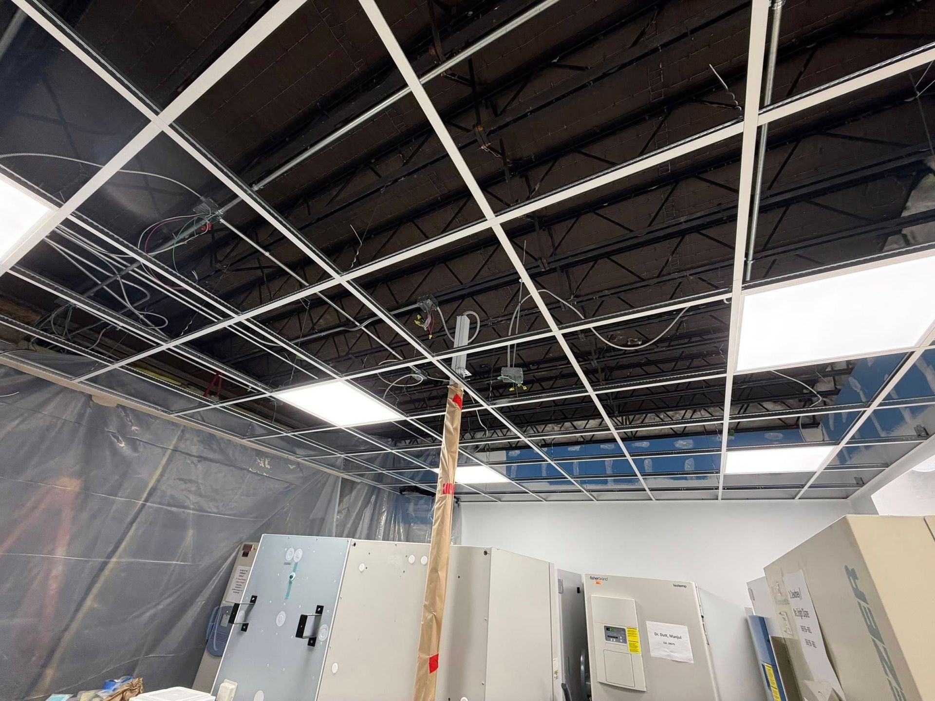 View of a drop ceiling under construction, with exposed black metal joists, wires, and light panels above plastic sheeting.